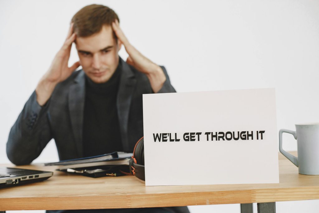A stressed man sits at a desk with a motivational sign saying 'We'll Get Through It'.